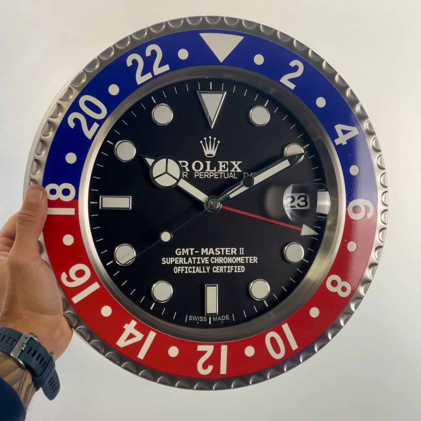 Hand holding a large wall clock designed like a Rolex GMT-Master II watch face with blue and red bezel, showing the time as 10:11:30 and date 23.